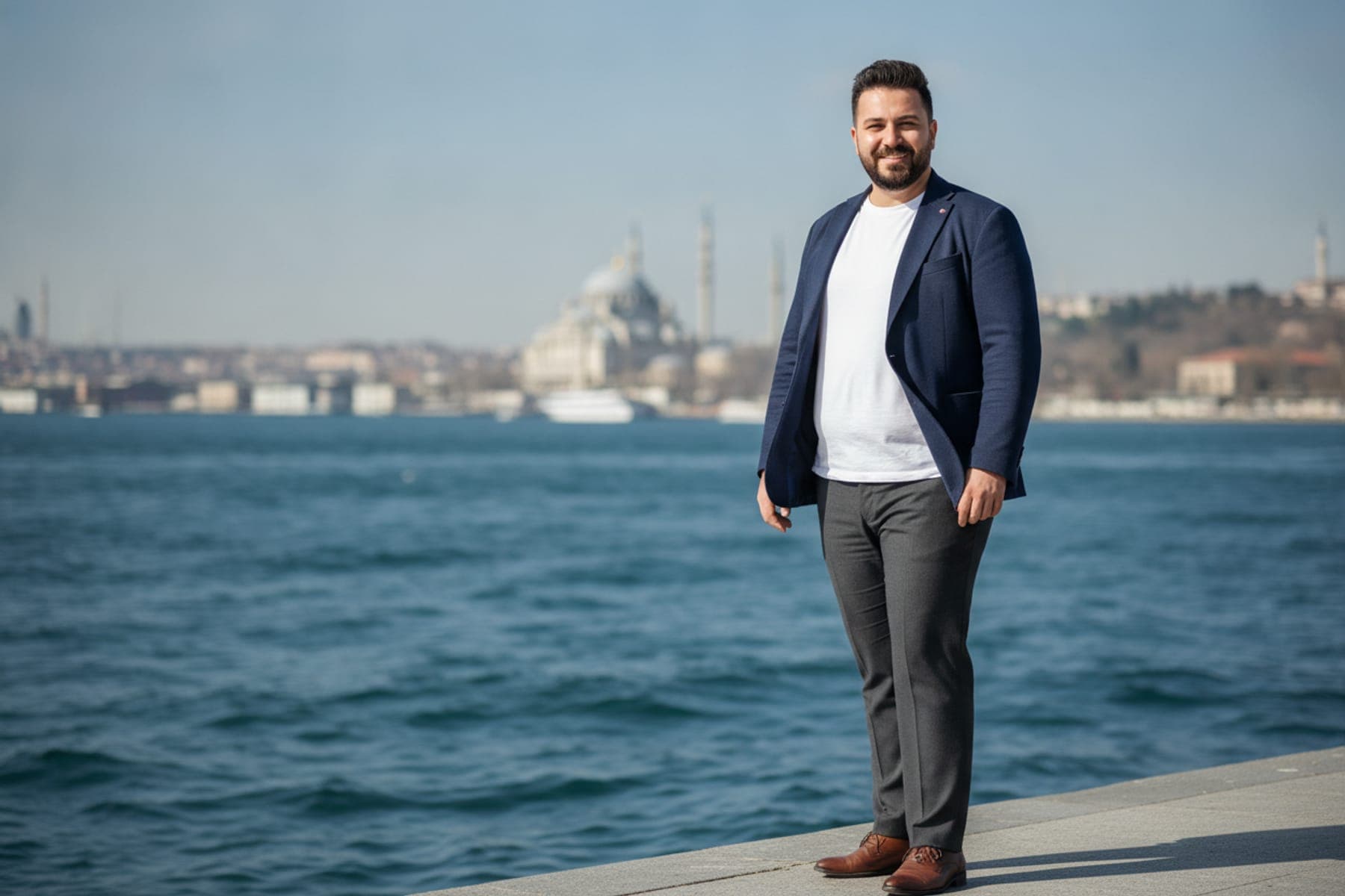 Büyük beden erkek smart casual kombini: lacivert ceket, beyaz tişört, gri kumaş pantolon ve kahverengi deri ayakkabıyla Boğaz kıyısında