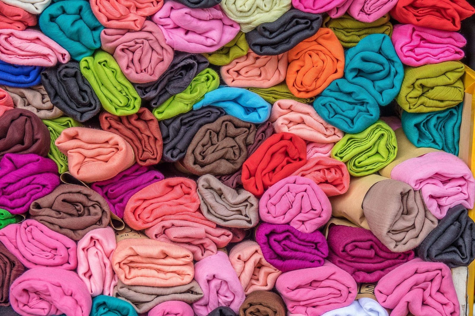 Farklı lif ve karışımlardan dokunmuş olabilecek pembe, turuncu, turkuaz ve gri rulo kumaşlar ve şallar